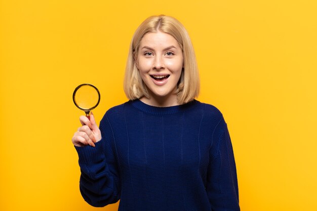 Blonde woman looking happy and pleasantly surprised, excited with a fascinated and shocked expression