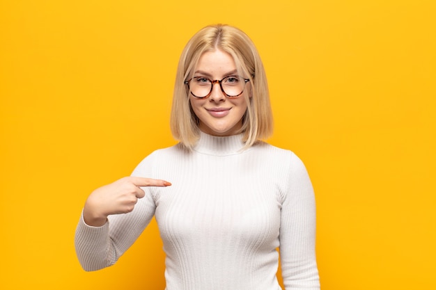 Blonde woman looking proud, confident and happy, smiling and pointing to self or making number one sign