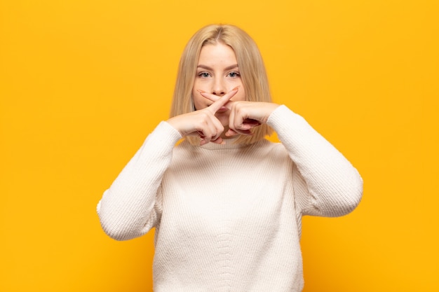 Blonde woman looking serious and displeased with both fingers crossed up front in rejection, asking for silence