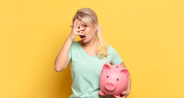 Blonde woman looking shocked, scared or terrified, covering face with hand and peeking between fingers