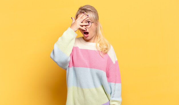 Blonde woman looking shocked, scared or terrified, covering face with hand and peeking between fingers