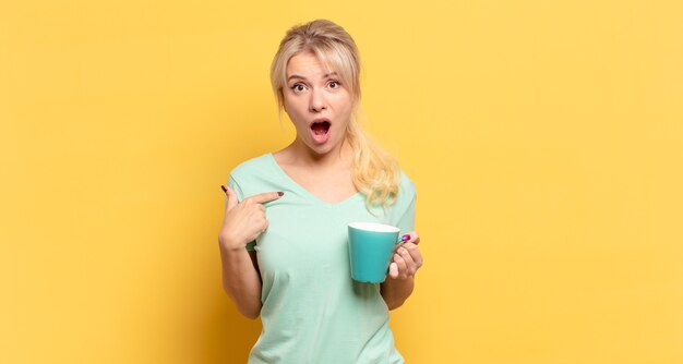 Blonde woman looking shocked and surprised with mouth wide open, pointing to self