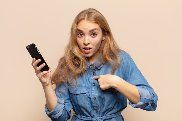 Blonde woman looking shocked and surprised with mouth wide open, pointing to self