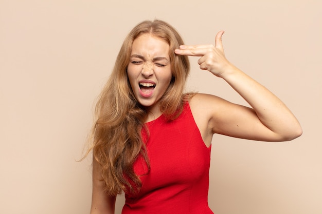 Blonde woman looking unhappy and stressed, suicide gesture making gun sign with hand, pointing to head