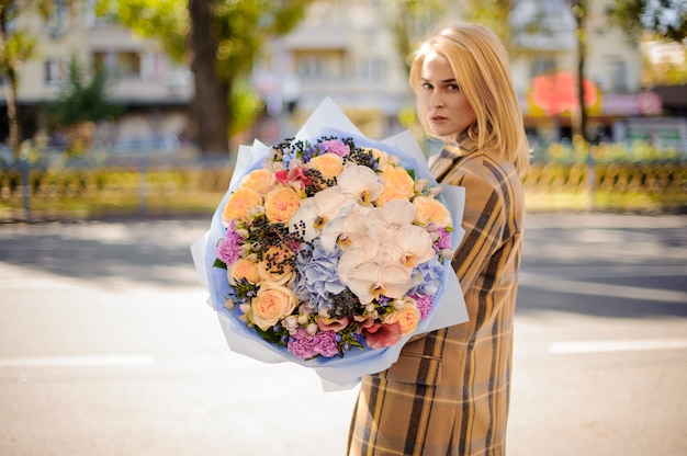 Donna bionda in cappotto scozzese che tiene un grande mazzo adorabile di fiori contro la città