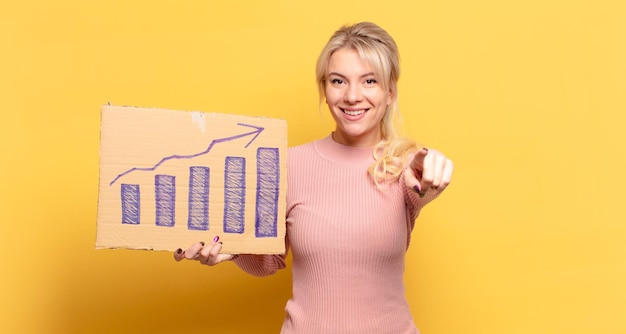 Blonde woman pointing at camera with a satisfied, confident, friendly smile, choosing you