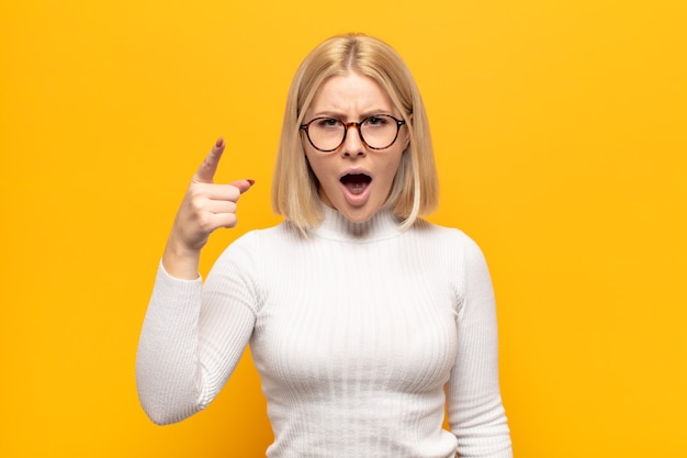 Blonde woman pointing at front with an angry aggressive expression looking like a furious, crazy boss