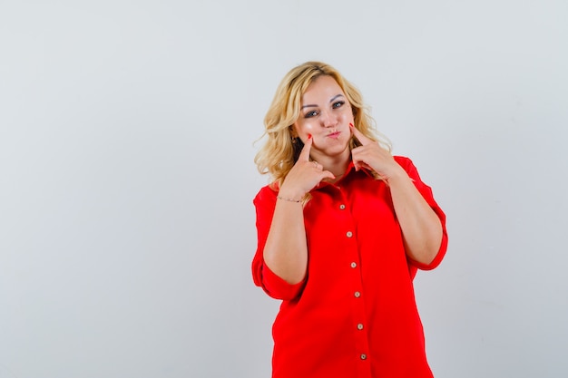 Blonde woman puffing cheeks, pointing to it with index fingers in red blouse and looking pretty. front view.