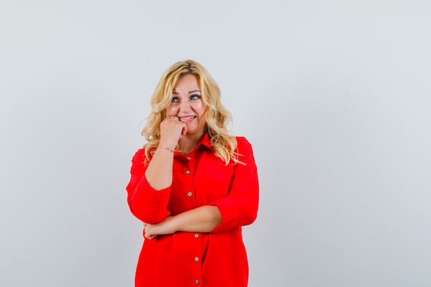 Blonde woman putting hand on cheek, standing in thinking pose in red blouse and looking pensive. front view.