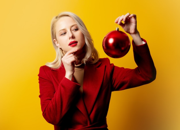 Blonde woman in red coat with Christmas bauble on yellow wall