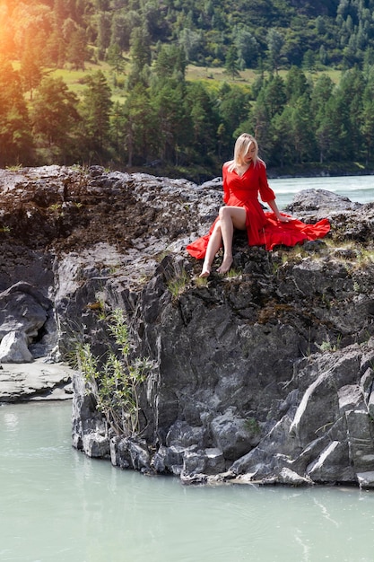 Blonde woman in red dress sits stretching her long legs on the rock stones on banks of Katun river against background of mountains on summer Freedom and vacation