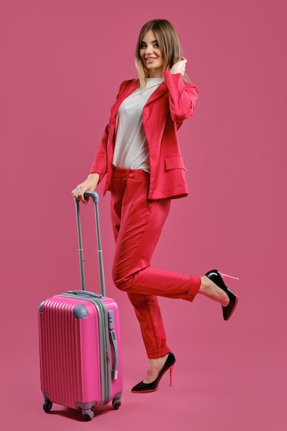 Blonde woman in red pantsuit white blouse high black heels she smiling posing with suitcase on pink