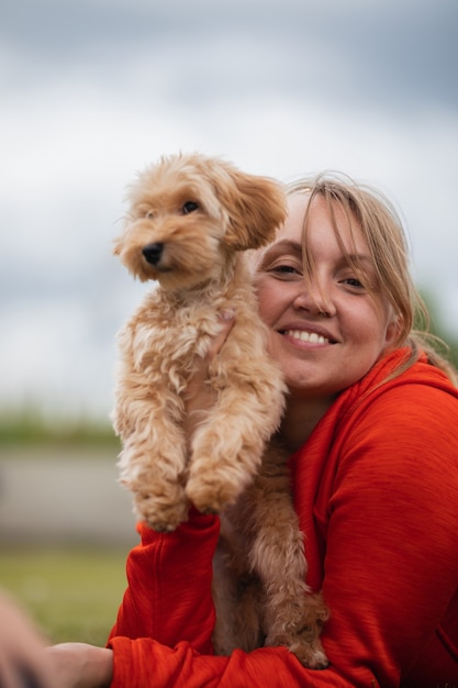 Blonde woman in a red sweater hugs a Maltipu dog outdoors