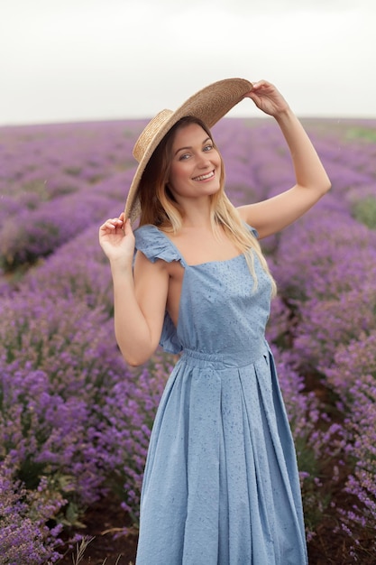 Blonde woman in round wicker hat and blue dress under the rain on the lavender field