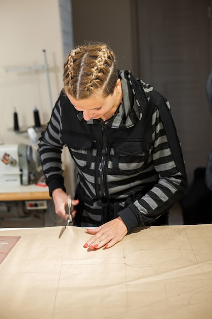金髪の女性の裁縫師は服を作るためのクラフト型紙からカットします。