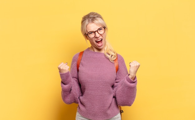 Blonde woman shouting aggressively with an angry expression or with fists clenched celebrating success