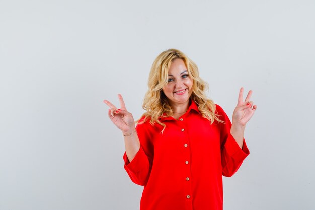 Blonde woman showing peace gestures with both hands in red blouse and looking happy , front view.