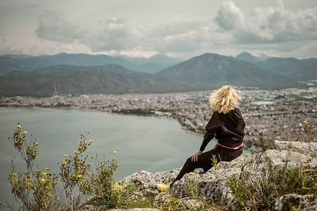 Una donna bionda siede su un'alta roccia che domina la città di mare e la baia del mar egeo in una giornata nuvolosa il concetto di viaggio e vacanza nella regione egea della turchia
