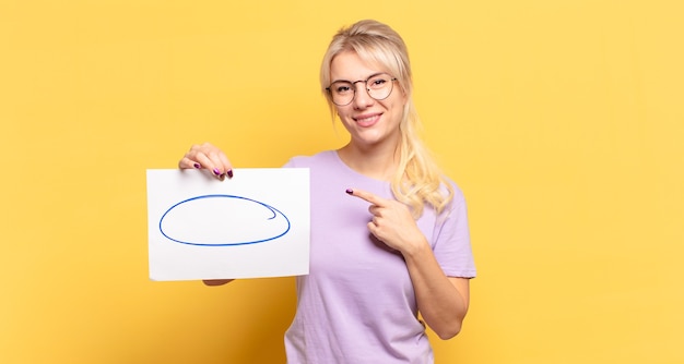 Blonde woman smiling cheerfully, feeling happy and pointing to the side and upwards, showing object in copy space