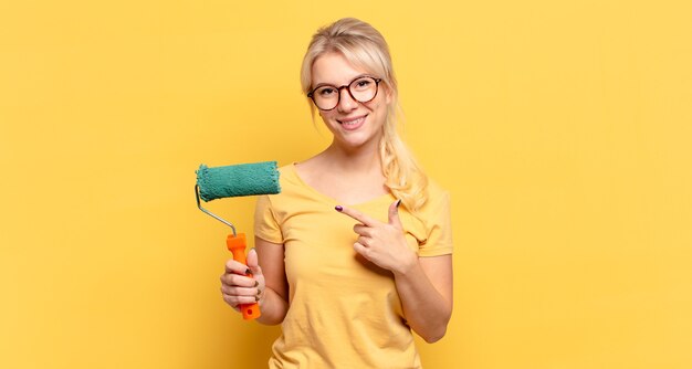 Blonde woman smiling cheerfully, feeling happy and pointing to the side and upwards, showing object in copy space