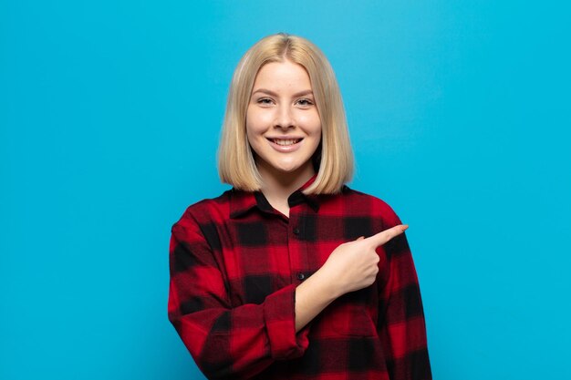 Blonde woman smiling cheerfully, feeling happy and pointing to the side and upwards, showing object in copy space