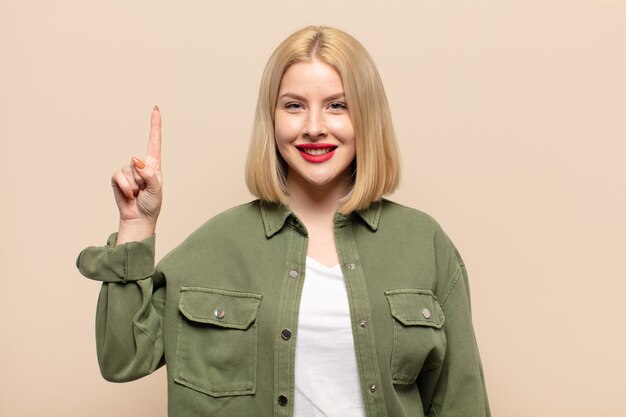 Blonde woman smiling cheerfully and happily, pointing upwards with one hand to copy space