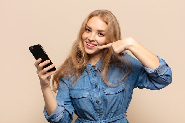 Blonde woman smiling confidently pointing to own broad smile, positive, relaxed, satisfied attitude