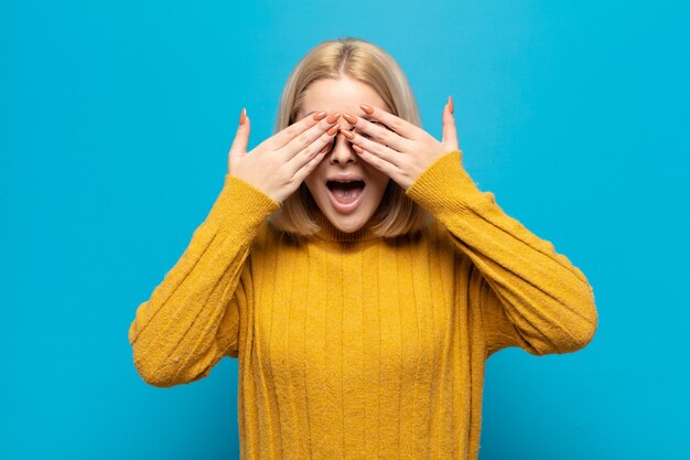 Blonde woman smiling and feeling happy, covering eyes with both hands and waiting for unbelievable surprise