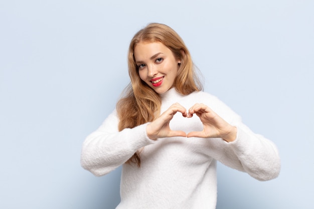 Blonde woman smiling and feeling happy, cute, romantic and in love, making heart shape with both hands