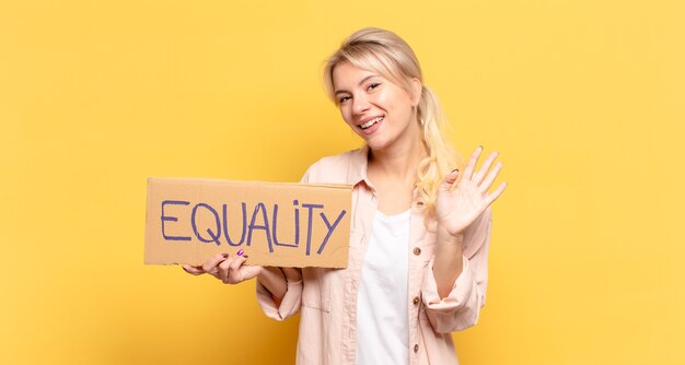 Blonde woman smiling happily and cheerfully, waving hand, welcoming and greeting you, or saying goodbye