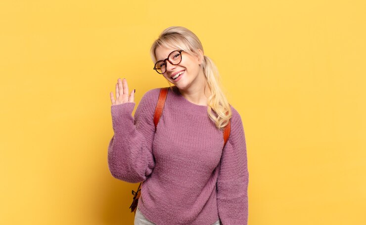 Photo blonde woman smiling happily and cheerfully, waving hand, welcoming and greeting you, or saying goodbye