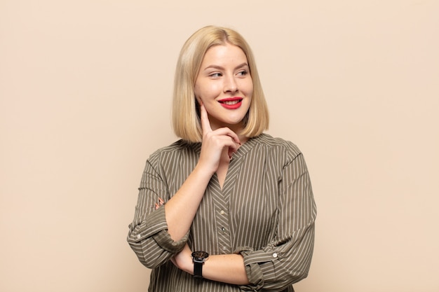 Blonde woman smiling happily and daydreaming or doubting, looking to the side