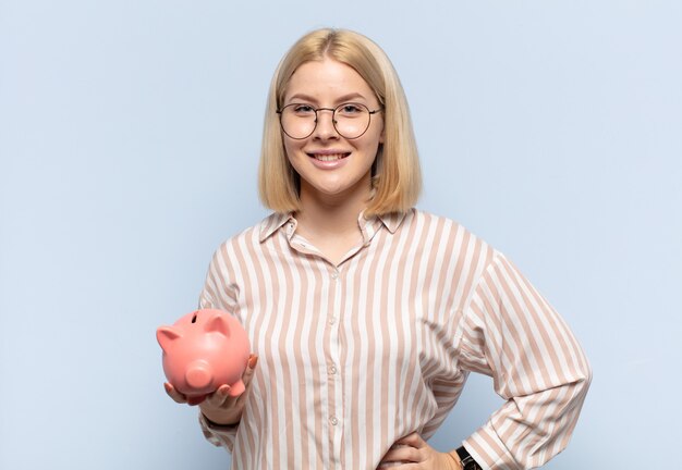 Blonde woman smiling happily with a hand on hip and confident, positive, proud and friendly attitude