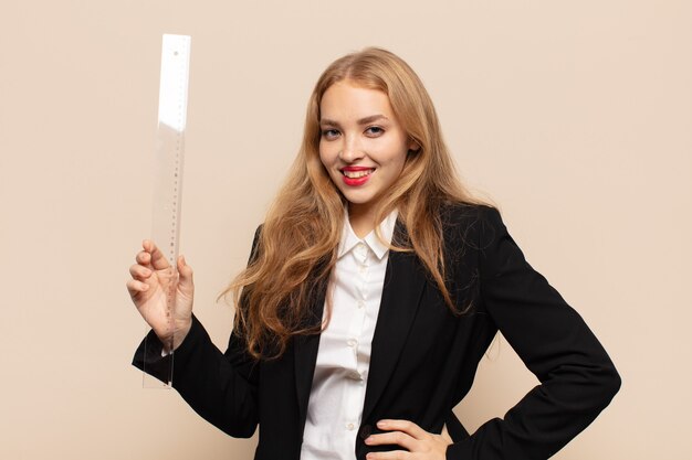 Blonde woman smiling happily with a hand on hip and confident, positive, proud and friendly attitude