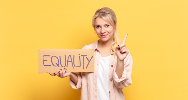 Blonde woman smiling and looking friendly, showing number one\
or first with hand forward, counting down