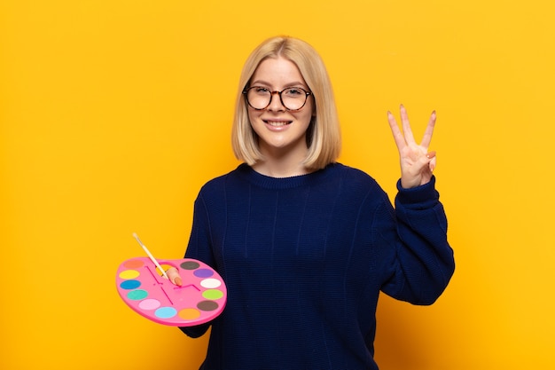 Blonde woman smiling and looking friendly, showing number three or third with hand forward, counting down