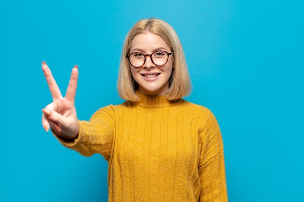 Blonde woman smiling and looking happy, carefree and positive, gesturing victory or peace with one hand