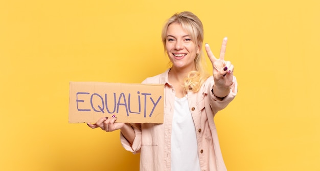 Blonde woman smiling and looking happy, carefree and positive,\
gesturing victory or peace with one hand