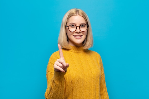 Blonde woman smiling proudly and confidently making number one pose triumphantly, feeling like a leader