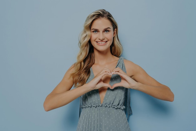 Blonde woman smiling showing heart gesture expressing love and affection isolated on blue background