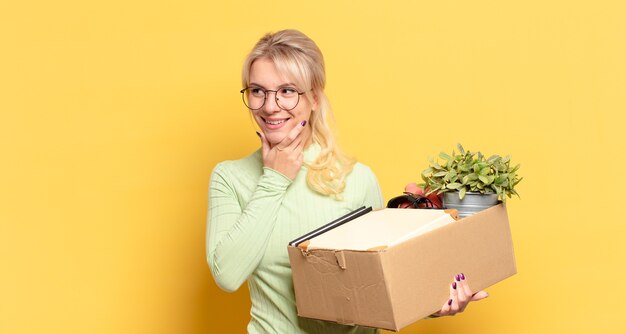 Blonde woman smiling with a happy, confident expression with hand on chin, wondering and looking to the side