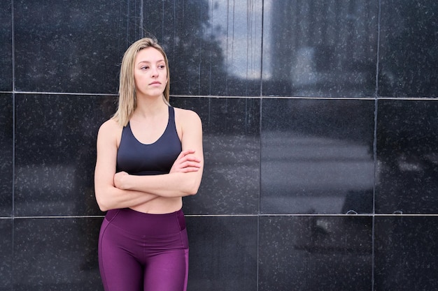 blonde woman in sportswear waiting on a dark wall with her arms crossed