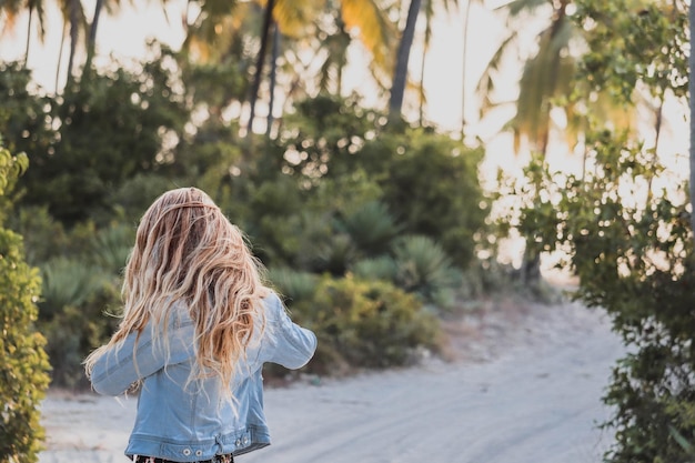 Blonde woman standing backwards with jeans jacket in magic tropic road