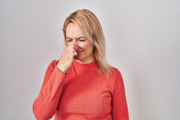 Blonde woman standing over isolated background smelling something stinky and disgusting intolerable smell holding breath with fingers on nose bad smell