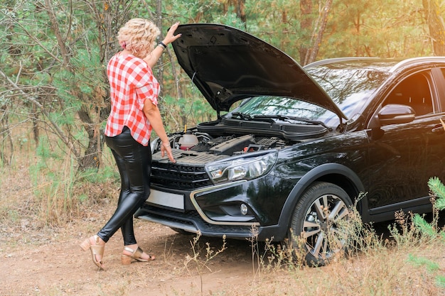 A blonde woman stands at the open hood of a car that broke down in the forest