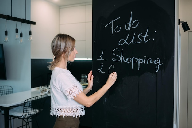 A blonde woman stands with her back and writes in chalk on a black board in the kitchen at home Todo list in the kitchen