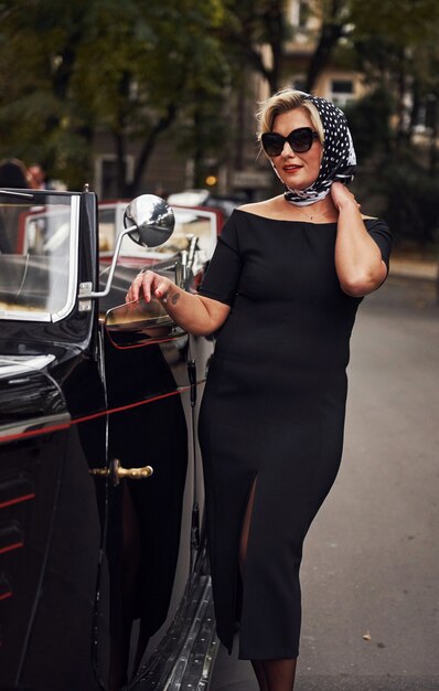 Blonde woman in sunglasses and in black dress near old vintage classic car.