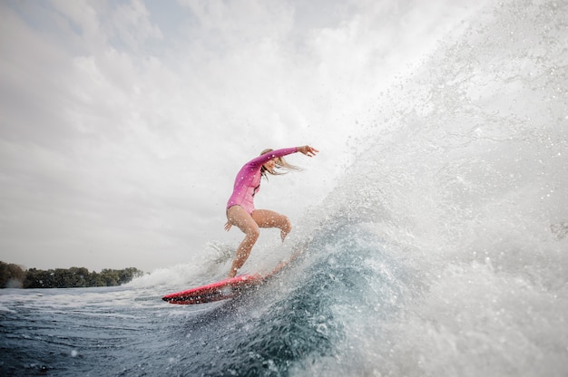 Surfista biondo della donna che guida giù l'onda di spruzzatura blu contro il cielo grigio