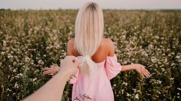 Blonde woman walking in the big endless field of daisies in summer evening. Lady wear pink dress