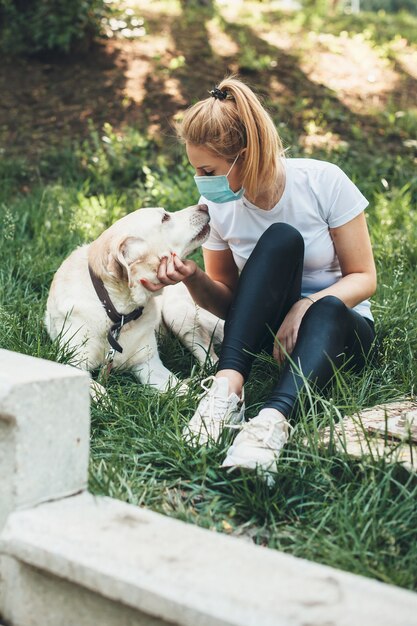 Blonde woman wearing an anti virus mask lying on the grass with\
her labrador